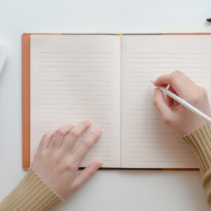 an open notebook with two hands resting upon it. One hand is holding it flat and the other hand is holding a pencil.