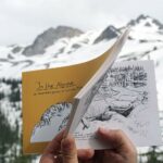 hands holding the open book "In the Alpine" with snowy mountain peaks above a treed slope in the background