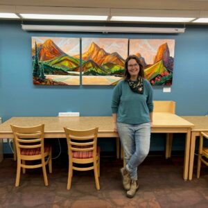 Artist Abby Wilson leans against wall next to a painting of a mountain.